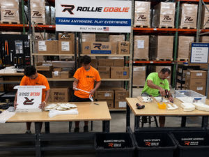 Three workers in orange and green shirts assemble or pack sports equipment at tables inside a warehouse, with Rollie Goalie branded signs and shelves of boxes in the background.